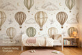Warm neutral hot air balloon wall mural spanning the full feature wall of a cosy toddler girl's bedroom — the panoramic beige and sage sky scene with white doves and gold moons above a low white toddler bed with cream linen and a blush knitted blanket.